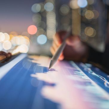Person using stylus on digital touch screen with city lights in background.