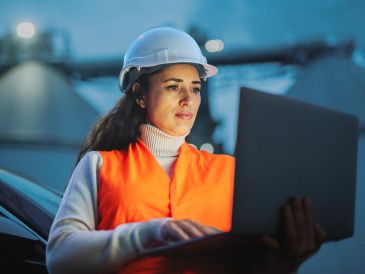Woman engineer wearing safety gear using a laptop at an industrial site.