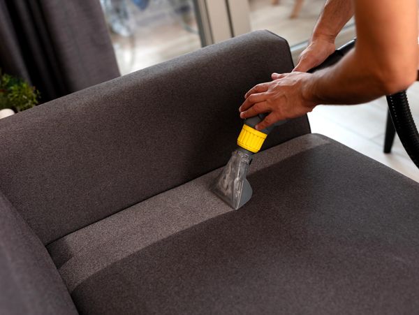 Person cleaning a gray sofa with a vacuum upholstery tool.