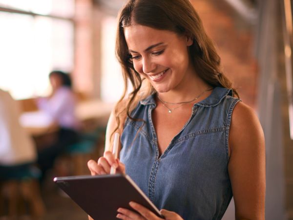 Smiling woman using a tablet with a stylus in a cozy café setting.