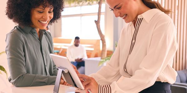 Two traveling sales women smiling and interacting at a reception desk with a tablet.