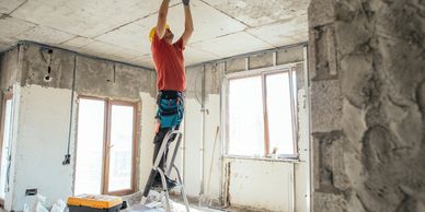 Worker in a hard hat fixing ceiling wires on a ladder inside a room under renovation.