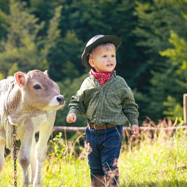 A young boy dressed as a cowboy stands next to a calf in a sunny field.