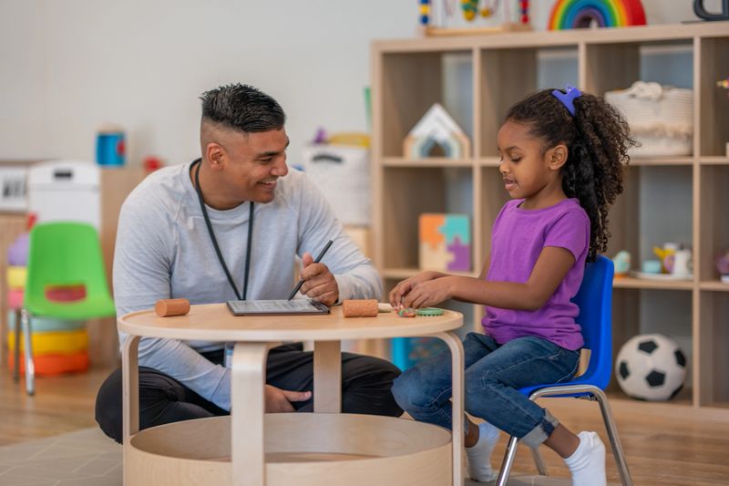 A male Occupational Therapist works with a young mixed race girl during a therapy session. He is using Play-Doh to work on the girls gross motor skills.