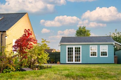 A blue garden shed with white-trimmed windows and doors in a backyard.