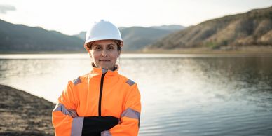 Female engineer in safety gear standing by a lake with mountains in the background.