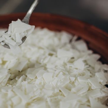 Close-up of white wax flakes in a wooden bowl with a spoon.