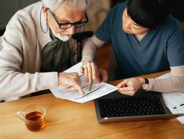 Caregiver assisting elderly man with paperwork at a table.
