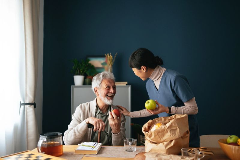 A smiling Japanese female caregiver bringing a paper bag with food, talking and laughing with a elderly patient who is sitting in the kitchen.
