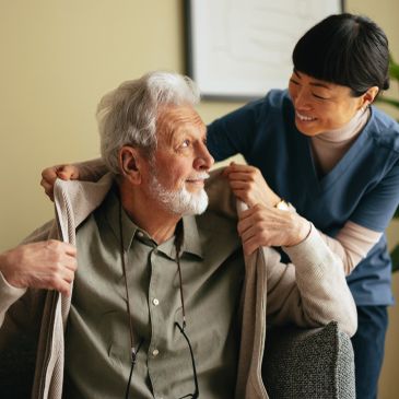 Caregiver helping elderly man put on a jacket with a smile.
