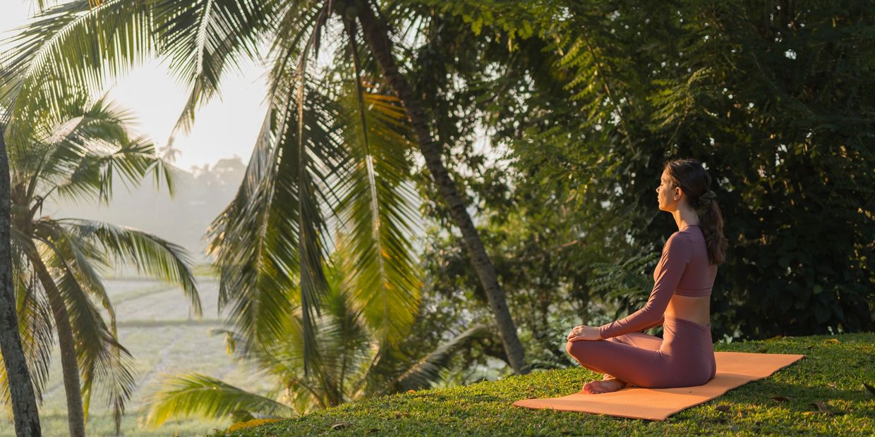 A woman meditates on a yoga mat in a lush, tropical setting at sunrise.