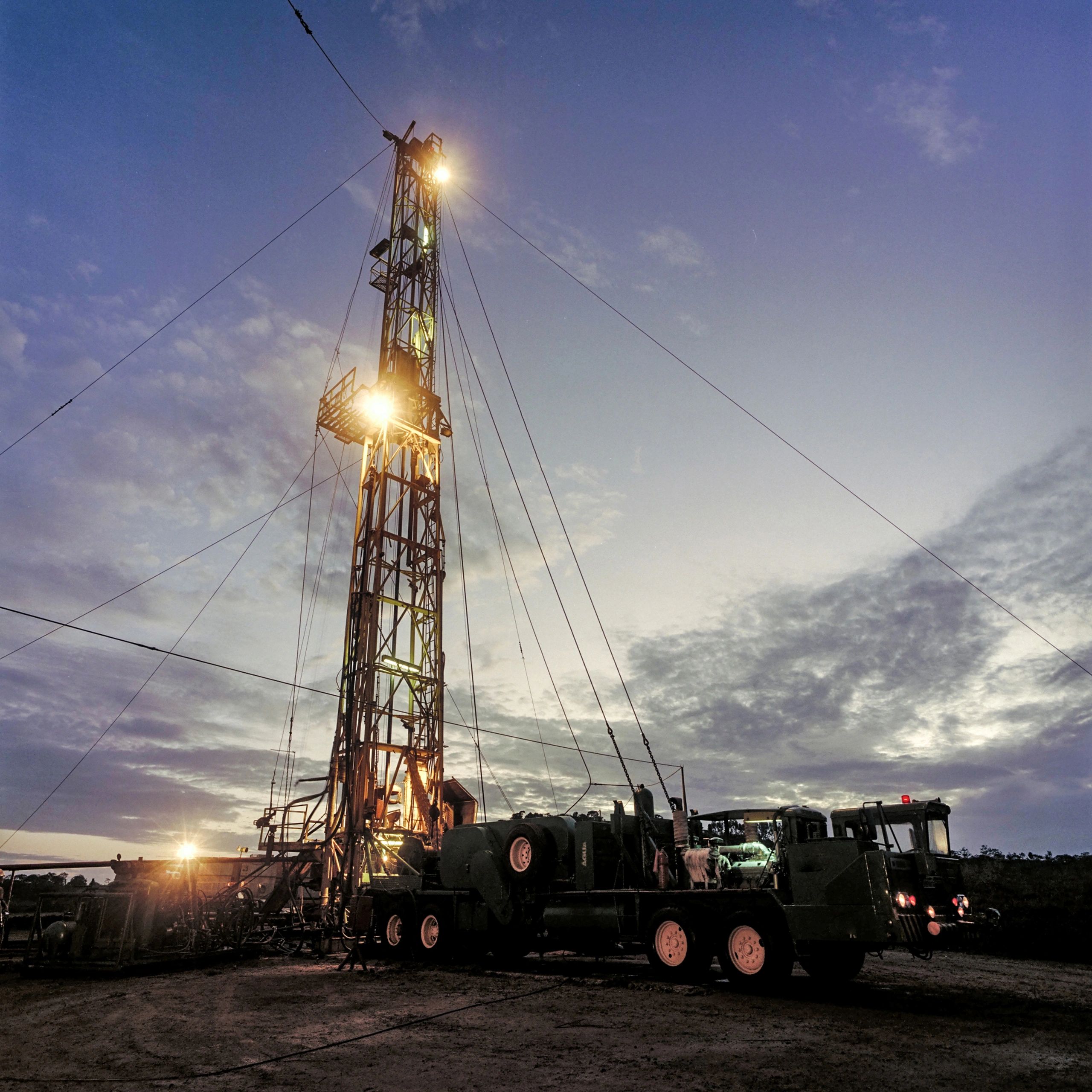 Illuminated oil drilling rig at dusk with attached machinery and equipment.