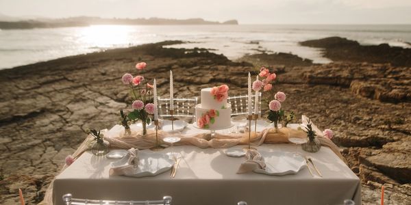 Elegant seaside table setup with floral decorations and a white cake at sunset.