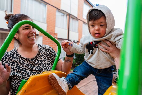 A woman helps a young child on a playground slide, both smiling and engaged.