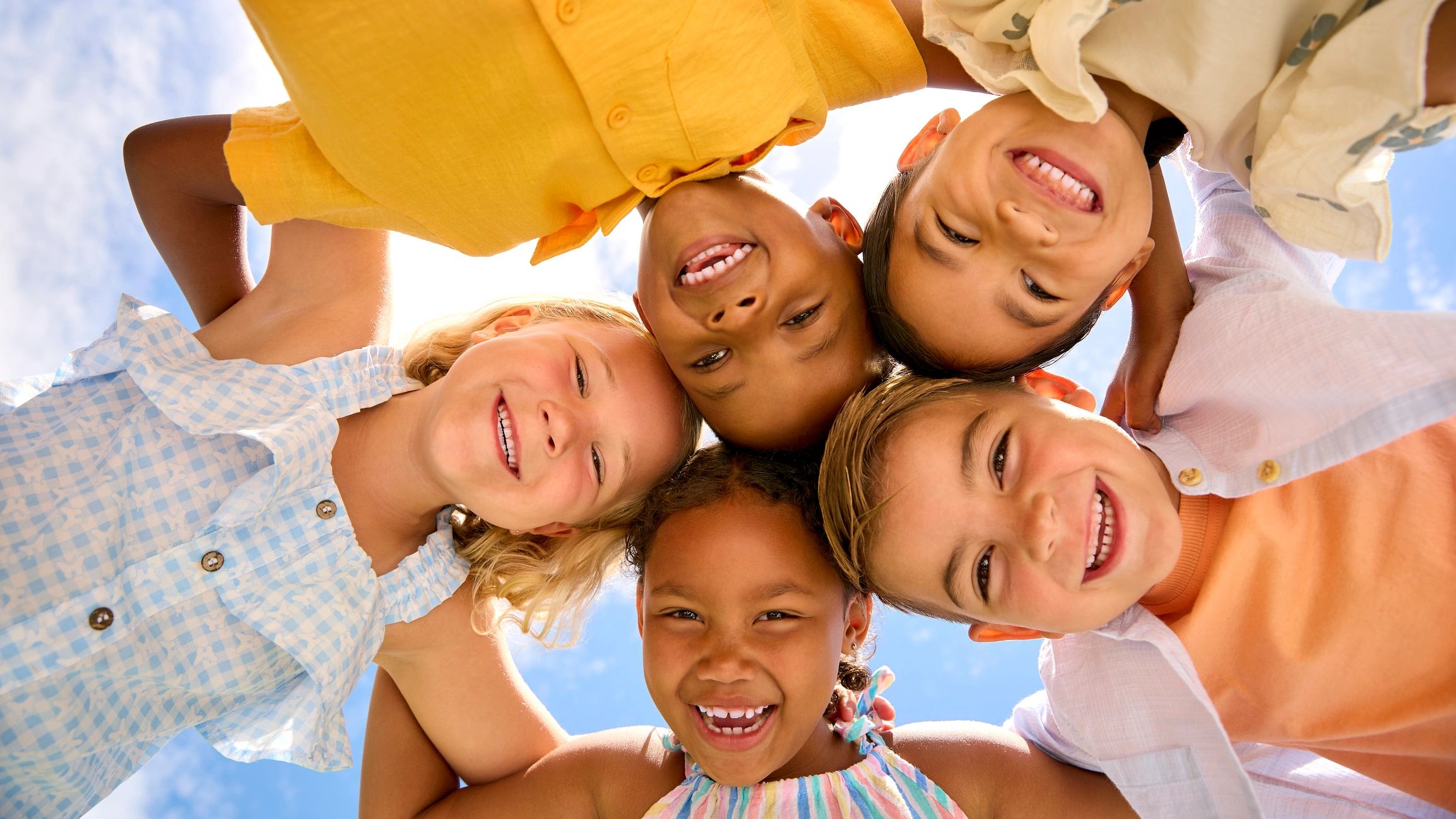 A group of five diverse children smiling together in a circle against a blue sky.