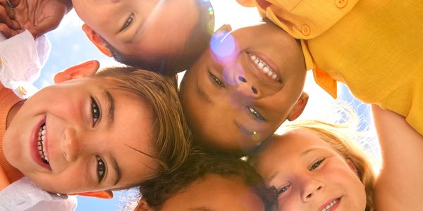 Five smiling children looking down at the camera in a bright outdoor setting.
