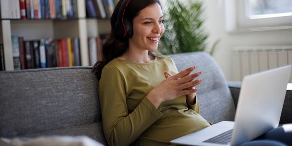 Pregnant woman wearing headphones and video chatting on a laptop at home.