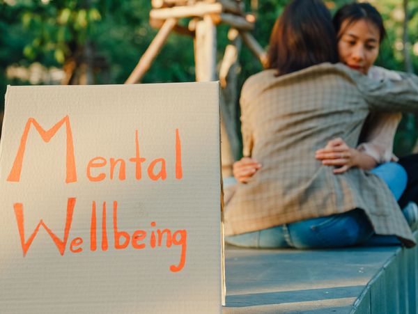 Cardboard sign reading 'Mental Wellbeing' with two people hugging in the background.