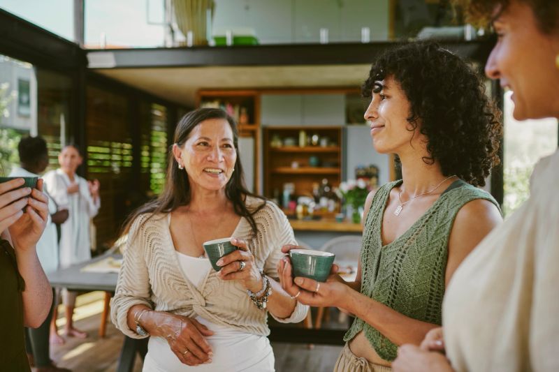 Diverse group of smiling female friends talking and drinking cups of herbal tea in the living room of a home during a wellness retreat