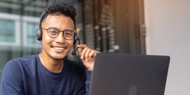 A man wearing glasses and a headset smiles while working on a laptop.