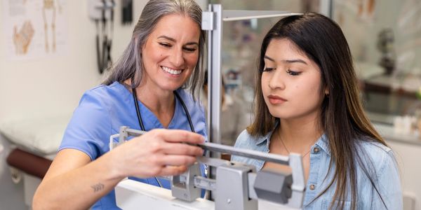 Nurse assisting a young woman with a weight measurement on a medical scale.