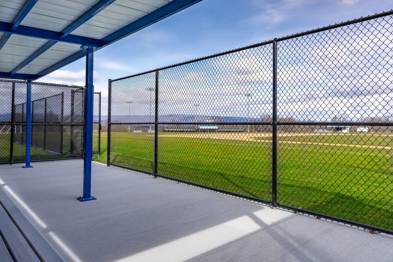View of typical nondescript high school baseball softball dugout with concrete floor, chain link fence, and blue roof shade. No people visible. Not a ticketed event.