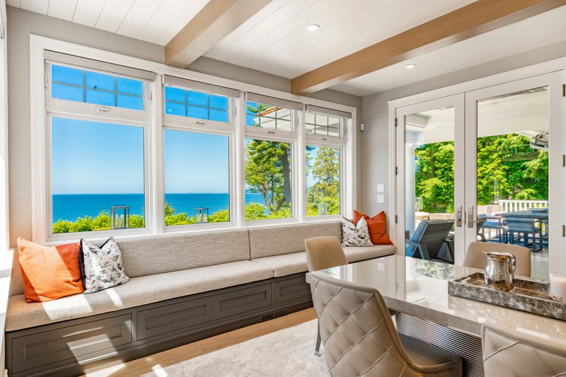 Photographs of the interior and exterior of a beautifully estate home in the Pacific Northwest, this home has been completely remodelled and is a waterfront home with vast views of the pacific ocean and san juan islands in the distance. Interiors show a grand entry foyer and staircase with wrought iron rails. Some interior views of the dining room with a banquette window seat looking over the sea and the patio contains outdoor furniture a fire pit and hot tub hot tub overlooking the grassy yard on a bright sunny day in July.