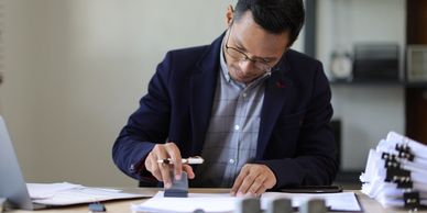 Man in glasses stamping a document at a desk with paperwork.