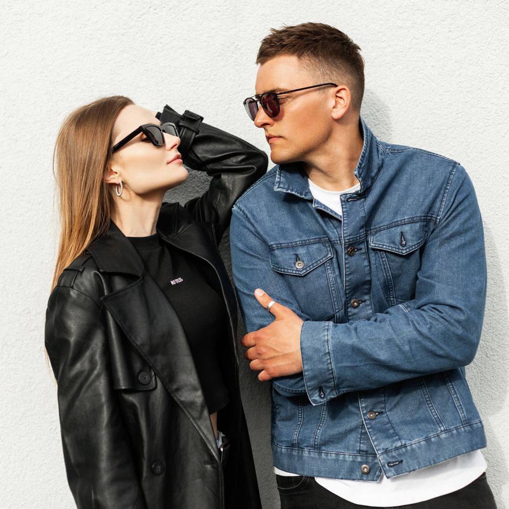 Stylish young man and woman in sunglasses pose confidently against a white wall.