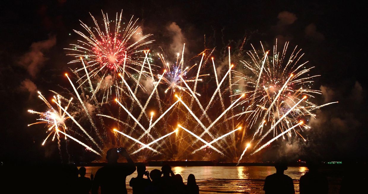 Spectacular fireworks light up the night sky over water, with silhouettes of spectators watching.