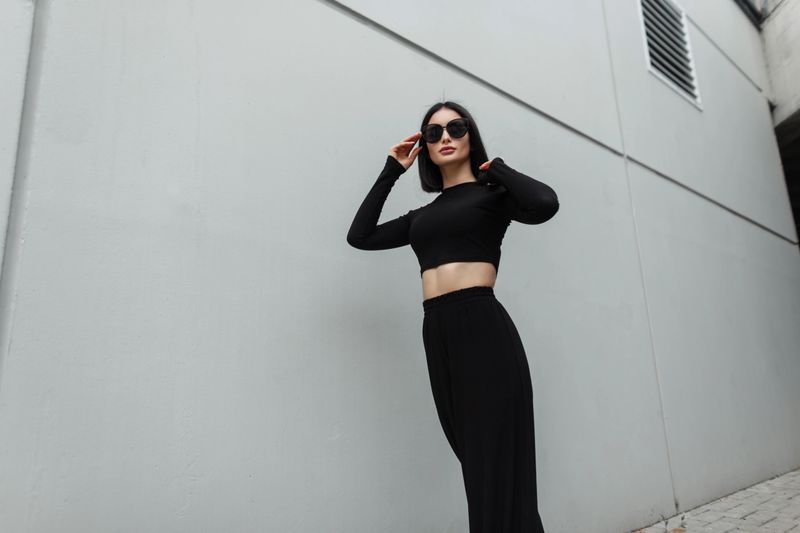 Fashionable pretty woman with fashion black sunglasses in stylish black outfit walks on the street near a gray building