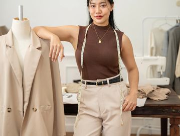 Fashion designer posing confidently with a measuring tape around her neck near a mannequin.