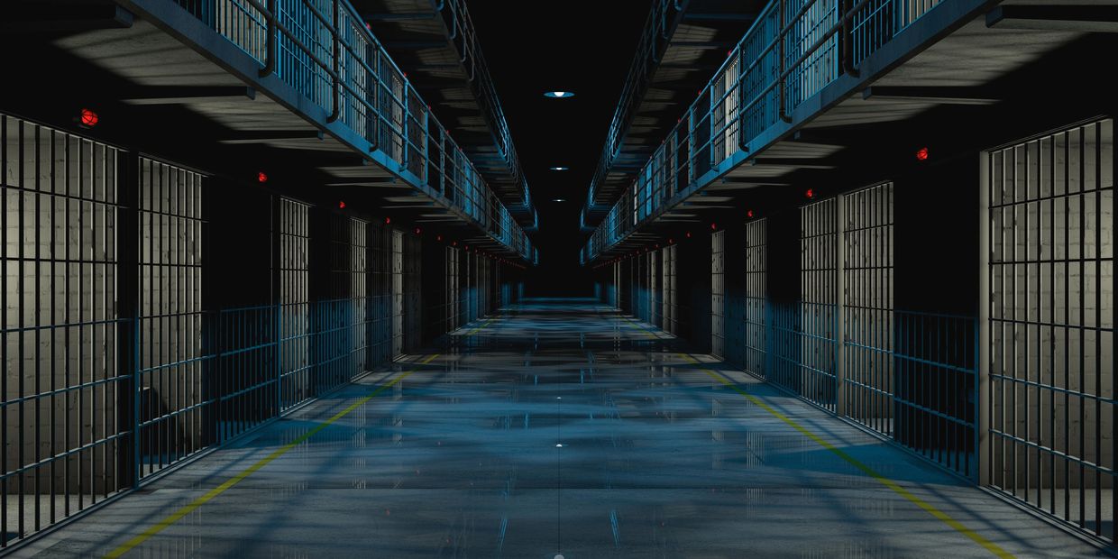 Dark, empty prison corridor with barred cells and blue railings.