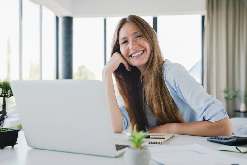 Creative young woman working in office laughing while using laptop at the desk. Female freelancer memorizing, creating ideas, making plans for future, dreaming at workplace