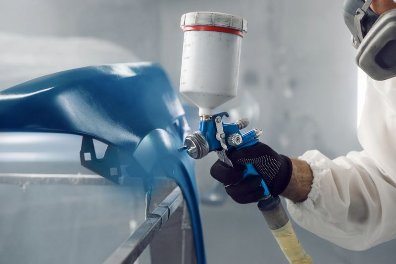 Airbrush coating. Automobile repairman painter in protective workwear and respirator painting car wing in paint chamber.