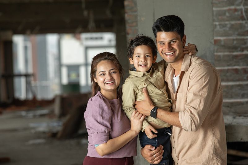 Portrait of cheerful young parents carrying cute son and standing in new apartment building under construction