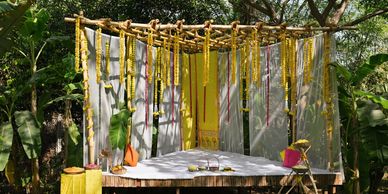 A decorated outdoor stage with yellow flower garlands and cushions among green plants.