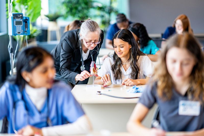A large group of medical students are seen sitting in rows of desk as they work in pairs on various activities.  They are each dressed professionally in scrubs and are working diligently on perfecting their skills.  Their professor is seen checking in on a student in the background.