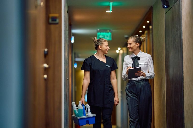 Happy housekeeping executive using touchpad while communicating with chambermaid in a hotel hallway. Copy space.