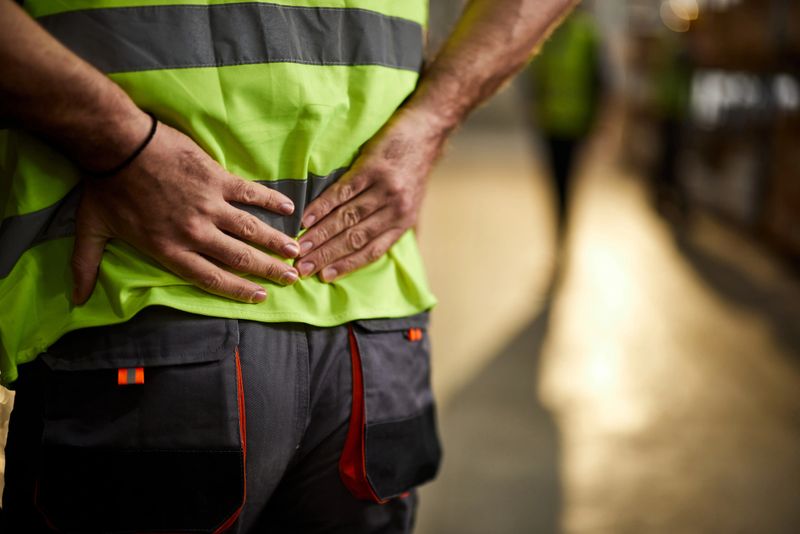 Close up of unrecognizable manual worker holding his back in pain at distribution warehouse. Copy space.