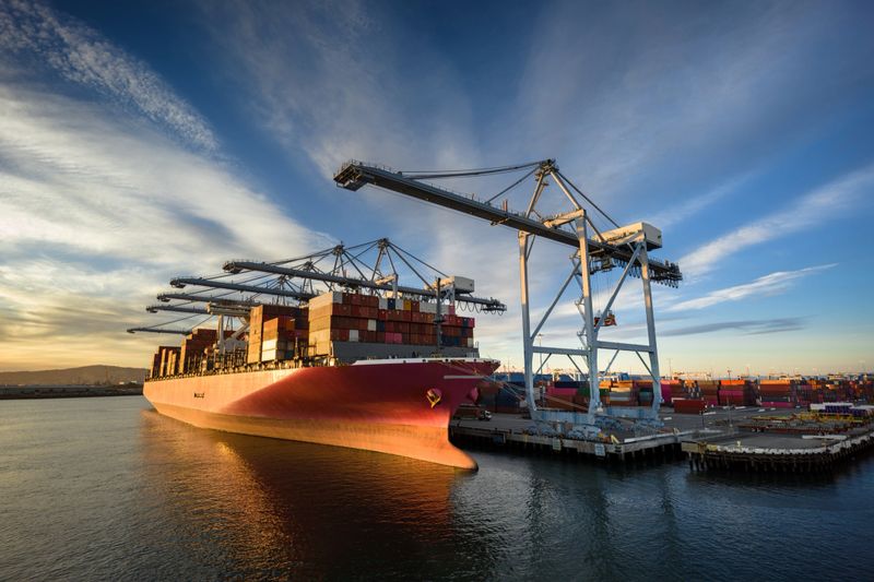 Aerial shot of a massive cargo ship loaded with containers docked in the Port of Long Beach, with the booms of ship-to-shore gantry cranes reaching out from the pier of the terminal.  

Authorization was obtained from the Port of Long Beach Security Division and POLB Harbor Patrol for this operation in restricted airspace.