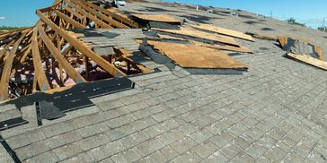 Damaged roof with missing shingles and exposed wooden beams under a clear blue sky.
