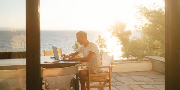 image of a man sitting at a table on a deck using his laptop while overlooking the ocean. 