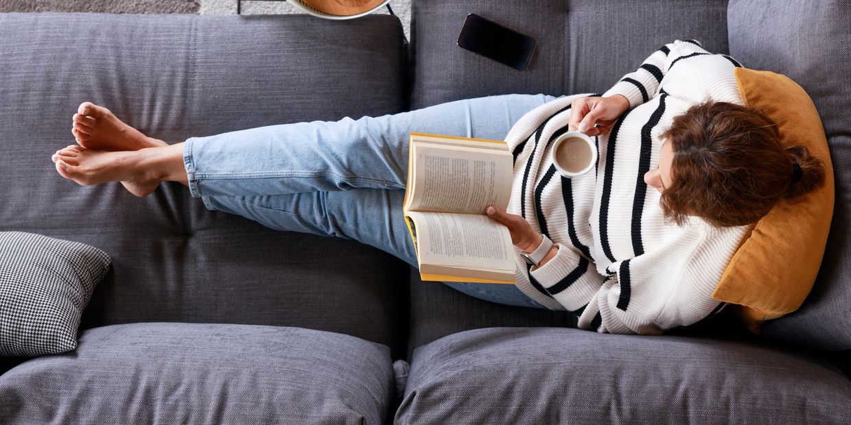 Person relaxing on a grey couch, reading a book and holding a cup of coffee.