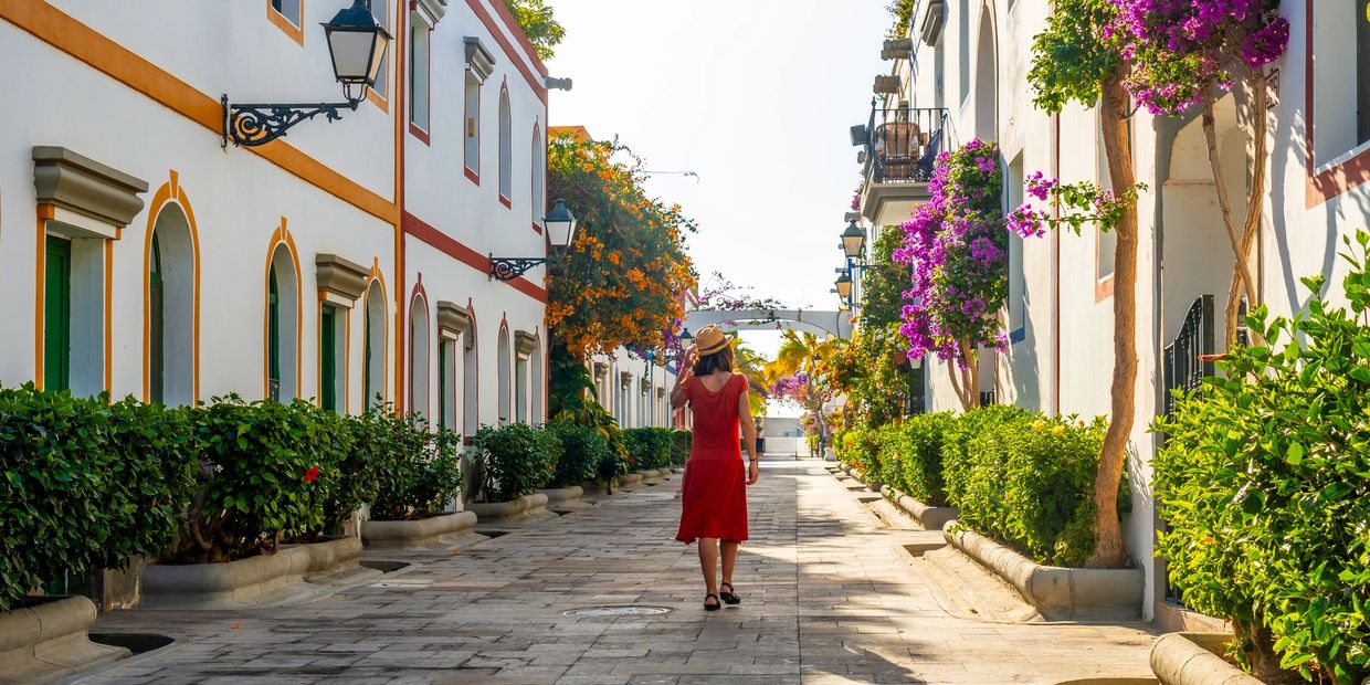 A woman in a red dress walks down a sunlit street lined with flowers and white buildings.
