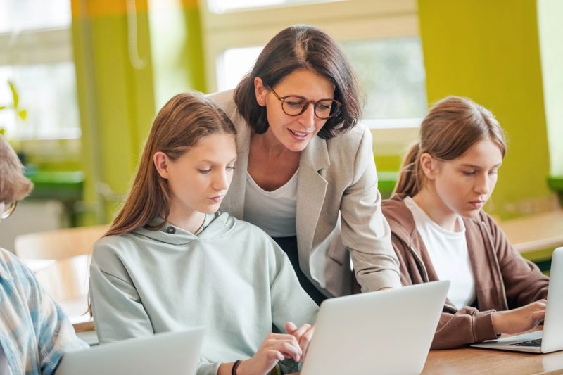 High school students sitting at the desk in the classroom during lesson, using laptops and talking with female teacher.