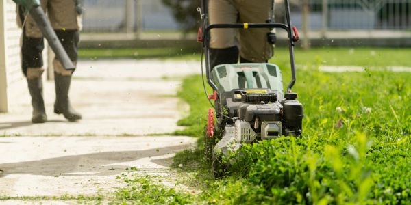 Person mowing a lawn with a blower operator in the background.