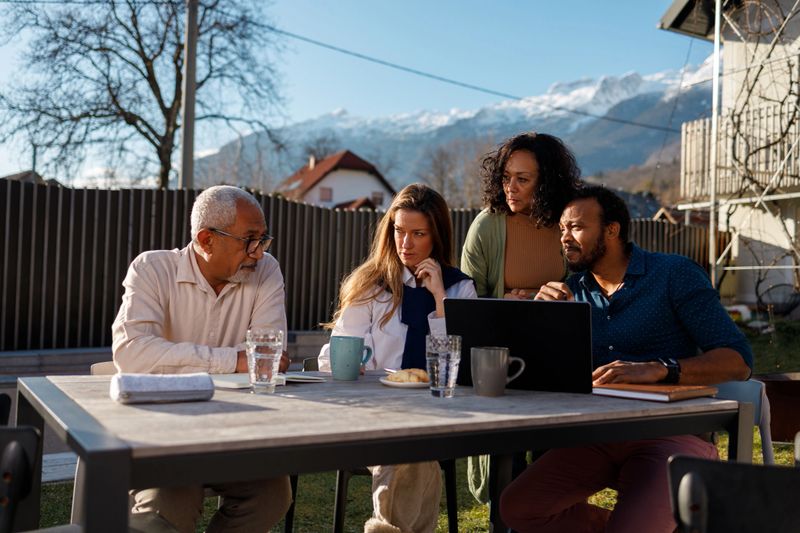 A family is gathered around a laptop outdoors, engaged in a serious business discussion, with a backdrop of mountains suggesting a balance between work and nature.