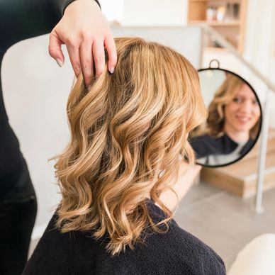 Woman with freshly styled blonde wavy hair looking in a round mirror at salon.