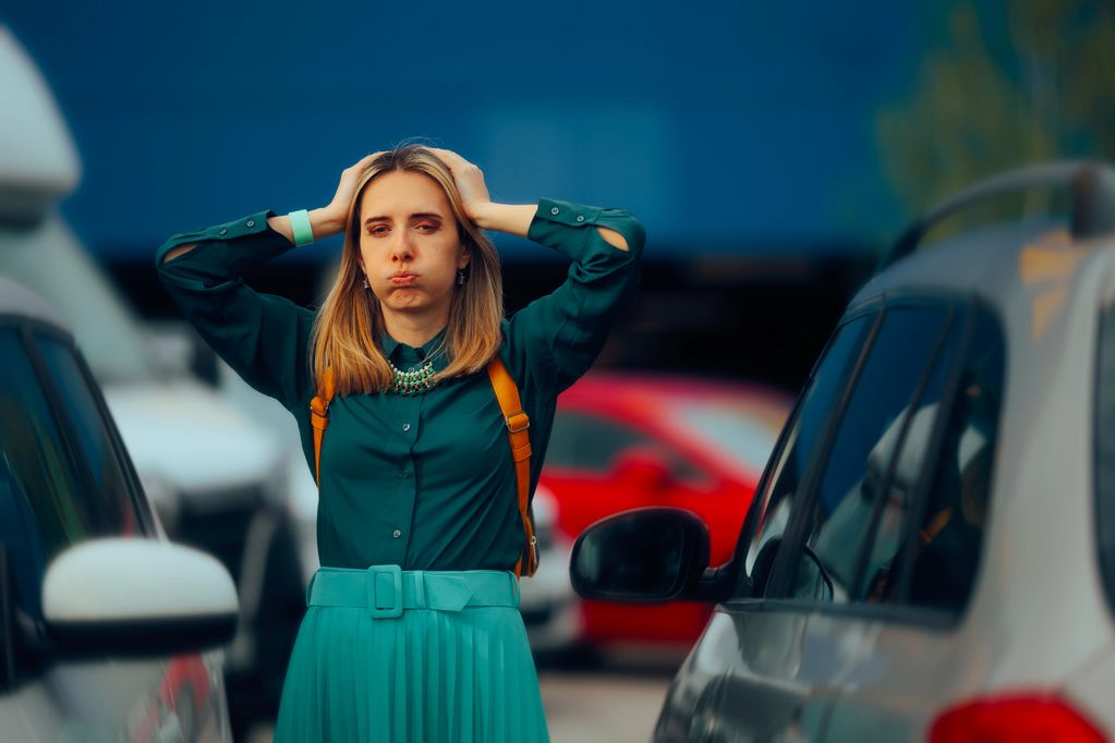 Frustrated woman standing between cars in a parking lot, holding her head.